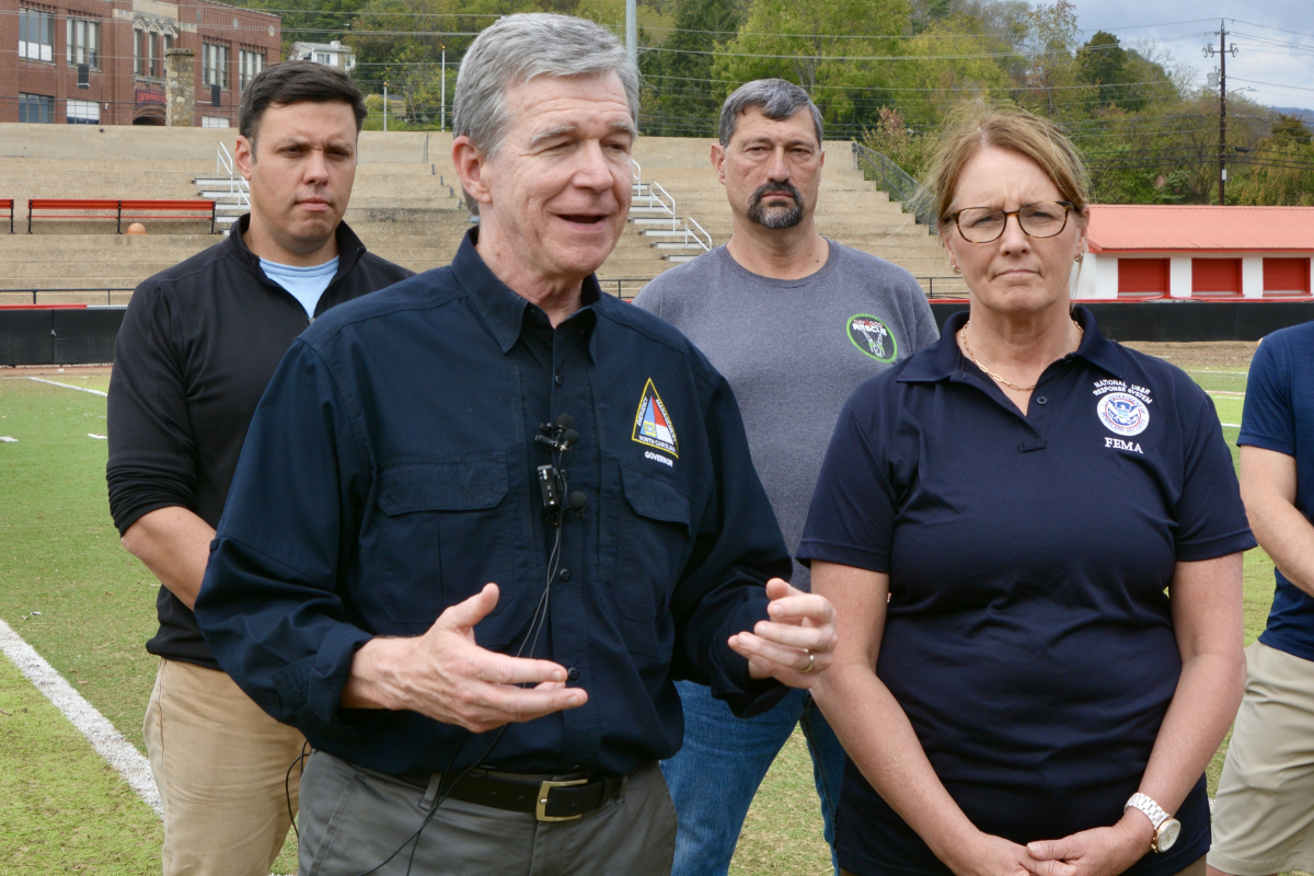 Canton Mayor Zeb Smathers (left) and Haywood Rep. Mark Pless (back right) look on as Gov. Roy Cooper and FEMA Administrator Deanne Criswell address reporters at Pisgah High School&#039;s Memorial Stadium.