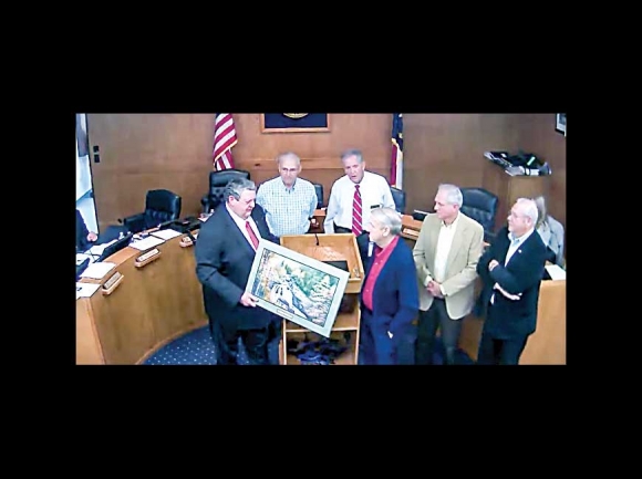 Macon County Commissioner Ronnie Beale (far left) presents a painting to Miles Gregory (center) in honor of his 25 years of service as the chairman of the Macon County Airport Authority. Screenshot